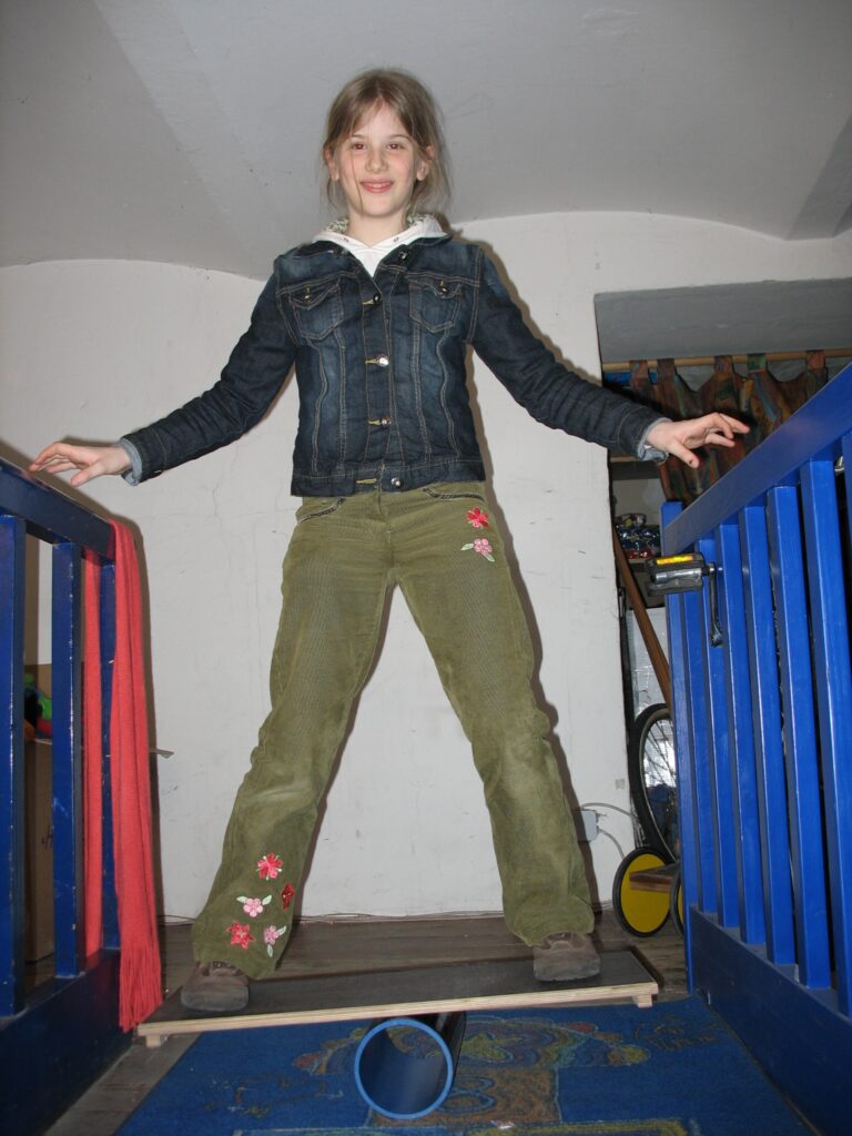 In Balance sein - zeigt ein Mädchen im Volksschulalter, das auf einem Holzbrett, welches auf einem Zylinder liegt, balanciert